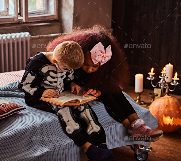 Three multiracial kids in scary costumes reading horror stories sitting on bed in an old house.