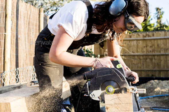Woman wearing protective goggles and ear protectors holding circular saw, cutting piece of wood on