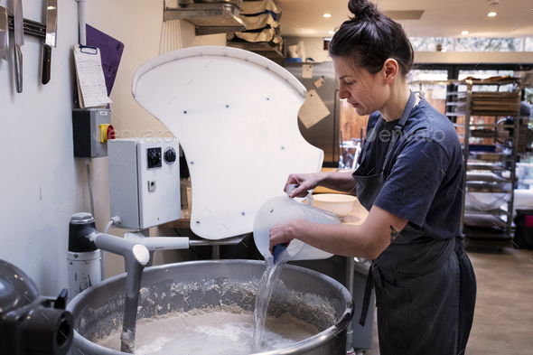 Woman wearing apron standing in an artisan bakery, pouring water into industrial mixer.