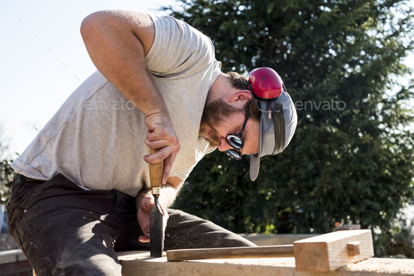 Man wearing baseball cap, sunglasses and ear protectors on building site, working on wooden beam.