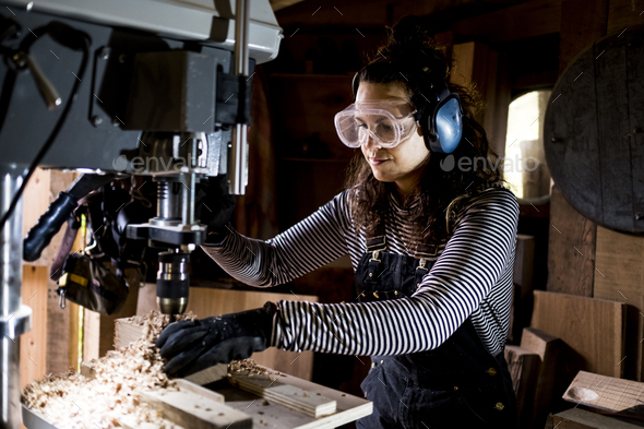 Woman with long brown hair wearing dungarees, safety glasses and ear protectors standing in wood