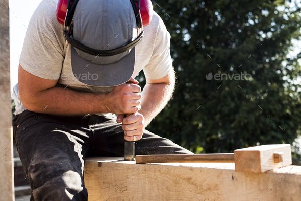 Man wearing baseball cap and ear protectors on building site, working on wooden beam.
