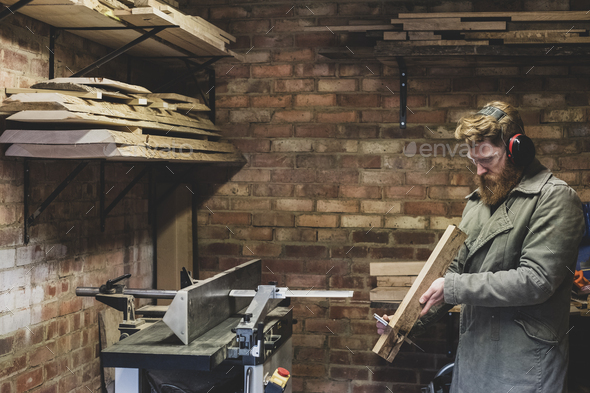 Bearded man standing in workshop, wearing ear protectors, working on piece of wood.