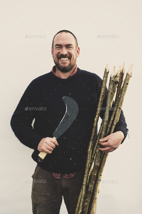 Bearded man holding bill hook and bunch of wooden stakes for traditional hedge laying, smiling at