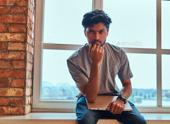 Pensive indian student holds the laptop while sitting on a window sill in a student dormitory.
