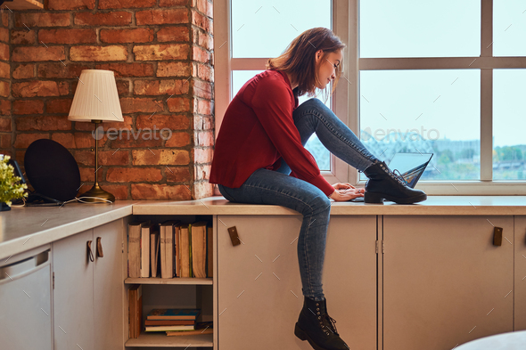 Young beautiful student girl sitting with laptop on window sill in student dormitory.