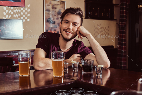 Young happy handsome guy resting in the bar or pub, sitting with glass of beer at wooden counter.