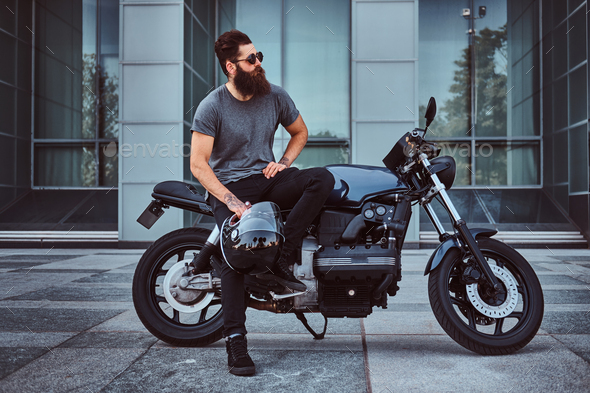 Brutal bearded male in a gray t-shirt and black pants sitting on his custom-made retro motorcycle