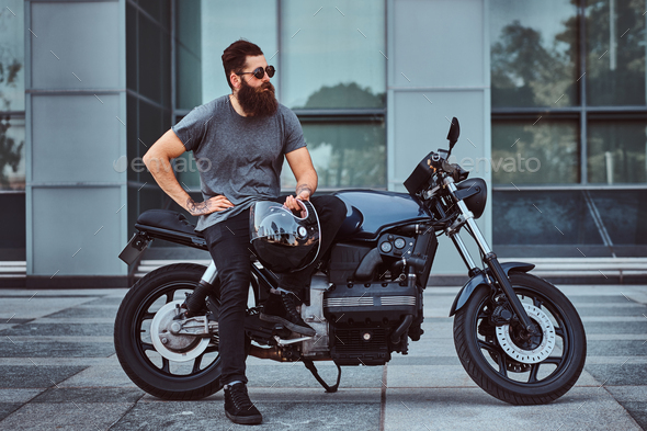 Brutal bearded male in a gray t-shirt and black pants sitting on his custom-made retro motorcycle