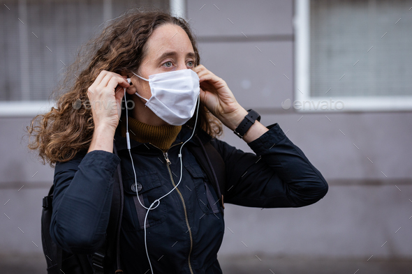 Caucasian woman putting on a protective mask in the streets, wearing earphones