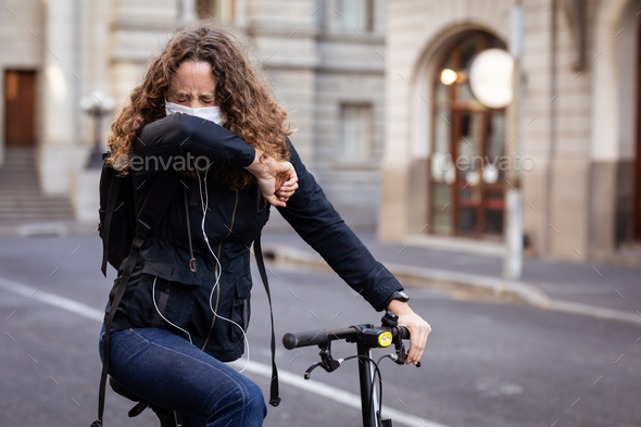 Caucasian woman wearing a protective mask on her bike, coughing and wearing earphones in the streets
