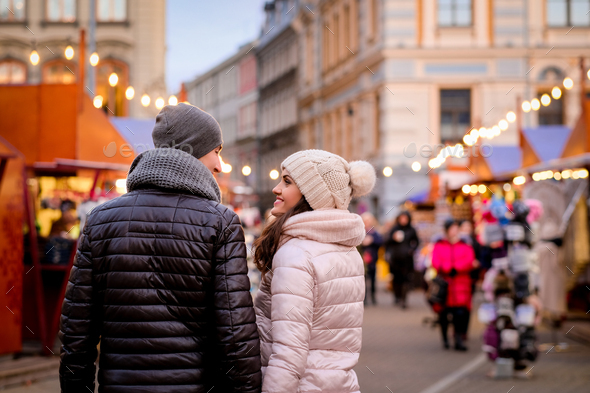 Romantic couple wearing winter clothes hugging while standing in evening street with Christmas fair