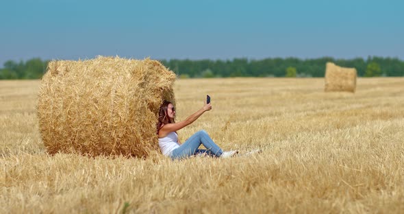 Young Woman Takes a Selfie in a Wheat Field She is Sitting By a Large Haystack alt