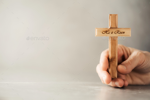 Hands holding wooden christian cross with text He is risen on grey background. Reminder of Jesus