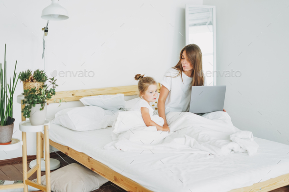 Mother freelancer with little daughter working on laptop in bed