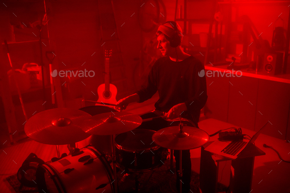 Young man in casualwear and headphones sitting by drumset in red lit garage