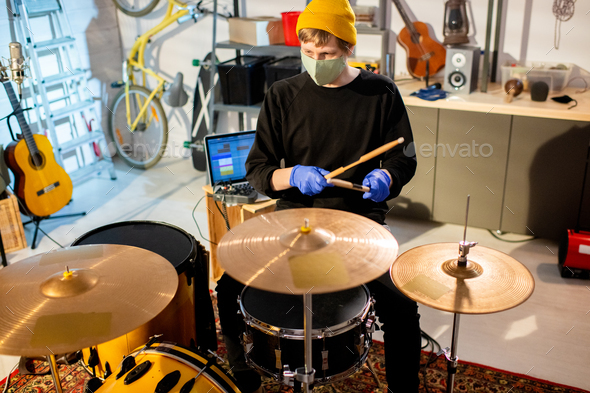 Young musician in protective gloves and mask sitting by drumset in garage
