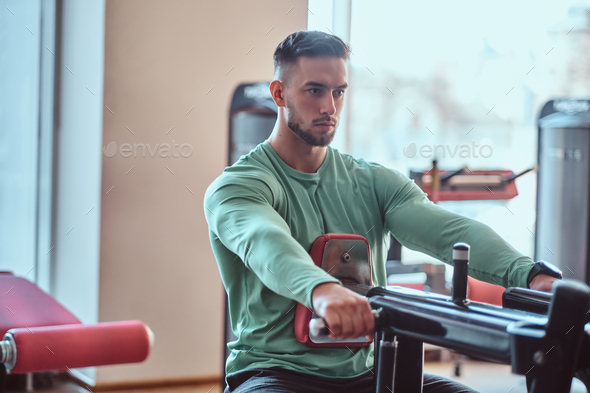 Young pensive bodybuilder in green shirt is doing exercises on training apparatus