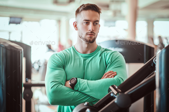 Young pensive bodybuilder in green shirt is standing near training apparatus with crossed hands