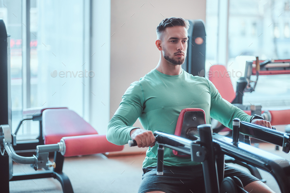 Young pensive bodybuilder in green shirt is doing exercises on training apparatus