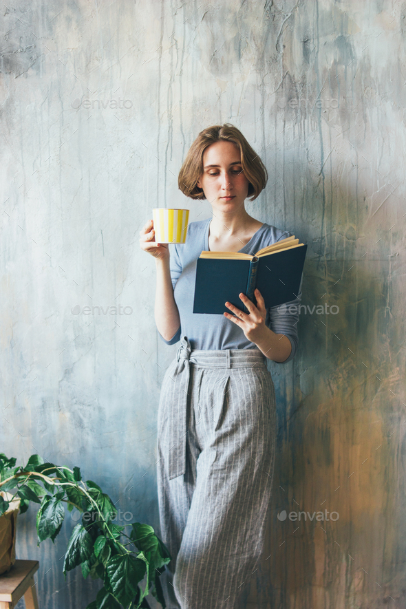 Young woman with yellow cup standing against wall and reading book in art work studio