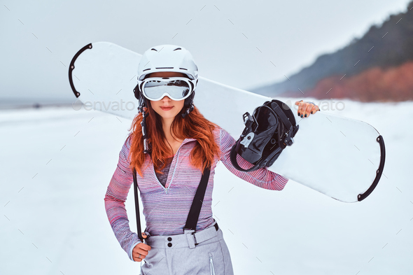 Beautiful girl in protective helmet and goggles holding a snowboard on a snowy Stock Photo by