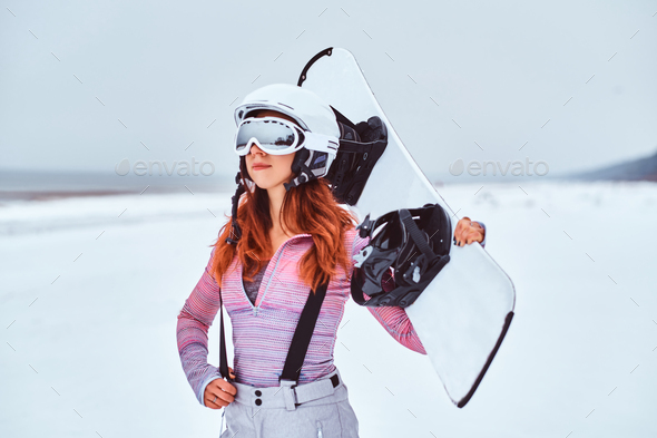 Beautiful girl in protective helmet and goggles holding a snowboard on a snowy Stock Photo by