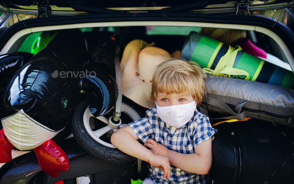 Small boy with face mask sitting in loaded car boot in countryside, local trip concept