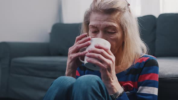 Lonely Old Woman Drinking Hot Beverage While Sitting on the Floor in Front of the Couch alt