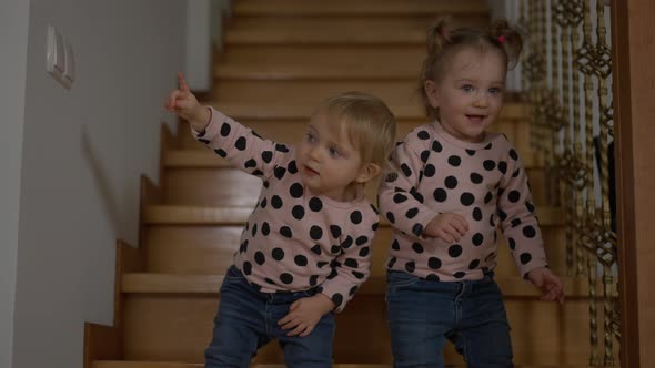 Beautiful Blond Twin Girls with Grey Eyes Standing on Stairs at Home Looking Away Smiling alt
