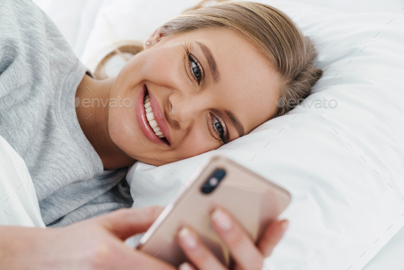Image of cute joyful woman using cellphone while lying after sleep