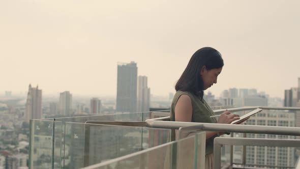 Woman using tablet chatting with friends while standing beside a balcony on a rooftop.