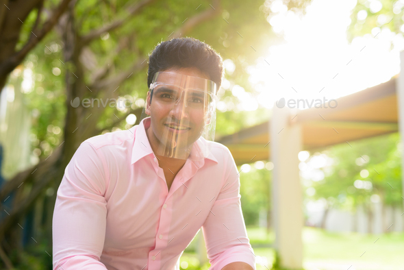 Happy young handsome Indian businessman with face shield smiling at the park - Stock Photo - Images