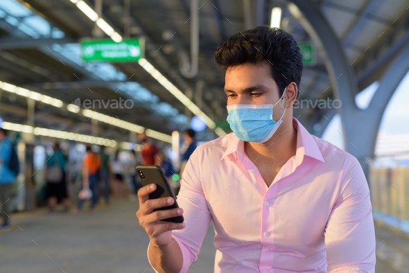 Young Indian businessman wearing mask and sitting with distance while using phone at the skytrain - Stock Photo - Images