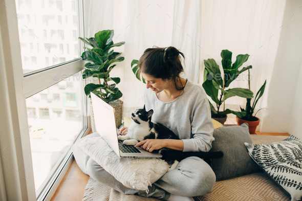 Casual girl working on laptop with her cat, sitting together in modern room with pillows and plants - Stock Photo - Images