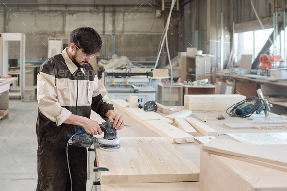 Young male worker in workwear and protective eyeglasses using grinding machine - Stock Photo - Images