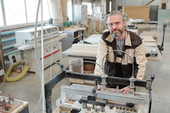 Senior worker of factory bending over industrial machine for processing wood - Stock Photo - Images