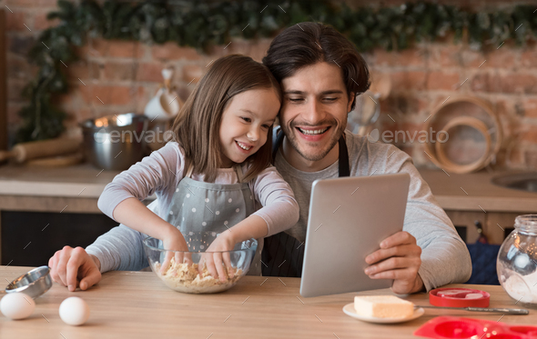Little Girl And Her Dad Checking Recipe On Digital Tablet While Baking