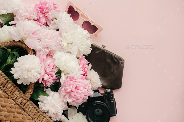 Stylish pink peonies in straw bag, photo camera, purse, retro sunglasses on pastel pink paper