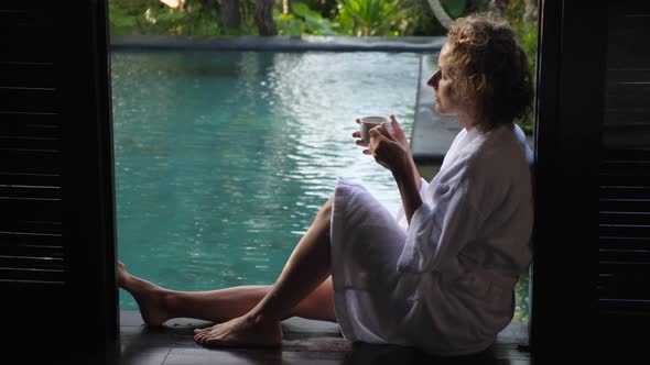 Side View of Woman Wearing Bathrobe Enjoying Her Morning Coffee Sitting on the Edge of the Swimming alt