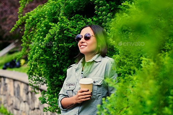 Brunette female in sunglasses drinks coffee in a summer park.