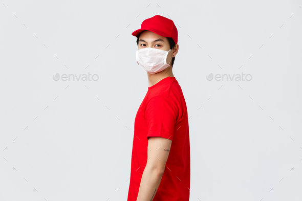 Portrait of asian delivery guy in red cap and t-shirt, wear protective medical mask, turn face to - Stock Photo - Images
