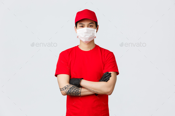 Confident young asian man in red delivery service uniform and cap, wearing protective medical mask - Stock Photo - Images