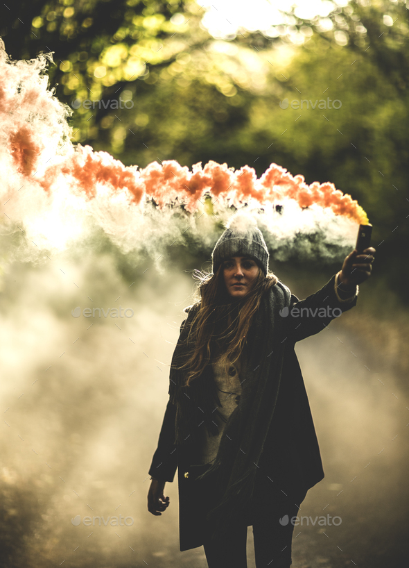 A young woman waving an orange smoke flare in a forest.