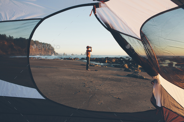 Man framed by camping tent, standing on beach and looking through binoculars at dusk, Olympic - Stock Photo - Images