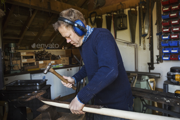 Man standing in a workshop, wearing ear protectors, holding a hammer, working on a piece of wood.