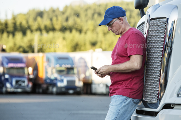 Caucasian man truck driver texting while standing next to the grill of his commercial truck in a