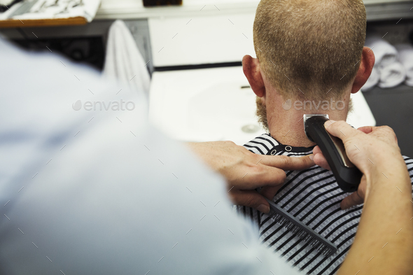 A customer sitting in the barber\'s chair, and a barber using a beard trimmer on his neck.