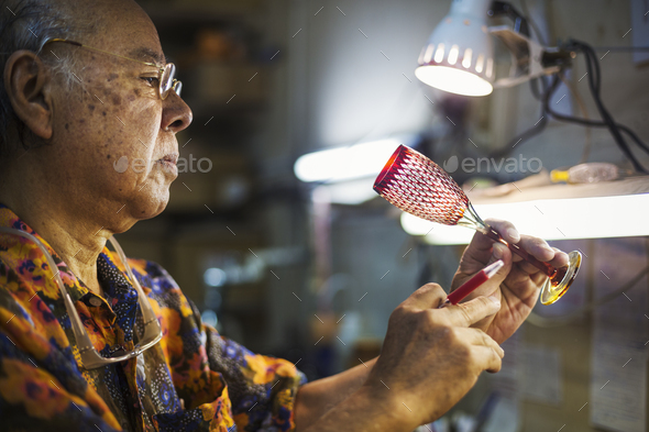 A senior craftsman at work in a glass maker's studio workshop, in inspecting red wine glass with cut