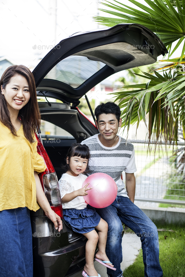 Portrait of Japanese woman, man and girl with pink ball sitting in open boot of car, smiling at